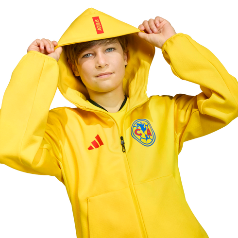 A boy wearing a yellow Adidas Club América hoodie with "ÁGUILAS" on the hood, holding its edges against a black background.