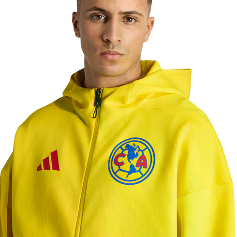 A man wears a yellow Adidas hoodie with the Club América crest against a black background.