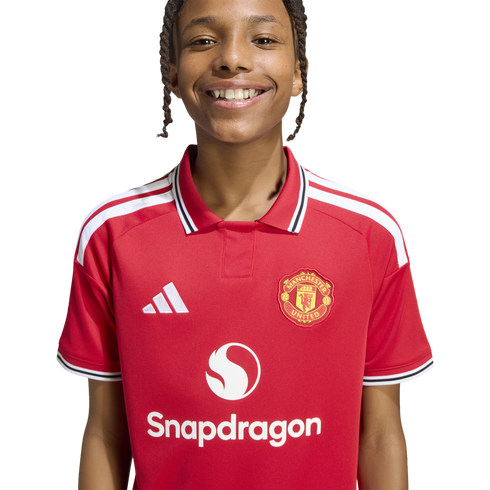 A smiling young person with braided hair wears a red Manchester United football shirt featuring the Snapdragon sponsor logo, set against a white background.