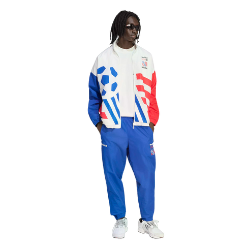 A person models a vintage-style Adidas World Cup USA '94 tracksuit against a black background. The white jacket features bold blue and red graphics, including a soccer ball and stripes, paired with matching blue track pants and white sneakers.