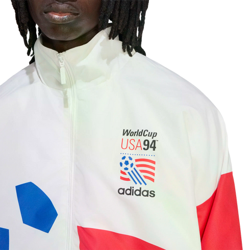 A close-up of a person wearing a white Adidas track jacket featuring the World Cup USA 94 logo on the left chest. The jacket has a high-neck zipper and red and blue color-blocked accents on the sleeves and body.