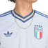 Close-up of a white Adidas Italy national team jersey featuring the FIGC crest, blue trefoil logo, and a blue and tan striped V-neck collar.