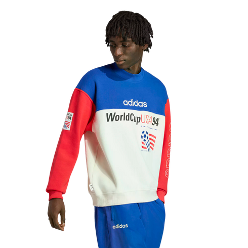 A person with short dreadlocks wearing an Adidas color-blocked "WorldCup USA 94" sweatshirt in blue, white, and red, paired with matching blue track pants, against a solid black background.