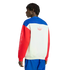 A rear view of a man wearing a color-blocked Adidas crewneck sweatshirt with blue, white, and red panels. The white back panel features a red-outlined Trefoil logo, and the left red sleeve has vertical "adidas" lettering in white. He is standing against a