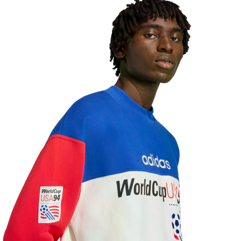 A close-up portrait of a man wearing a blue, white, and red color-blocked Adidas World Cup USA '94 sweatshirt against a solid black background.