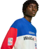 A close-up portrait of a man wearing a blue, white, and red color-blocked Adidas World Cup USA '94 sweatshirt against a solid black background.