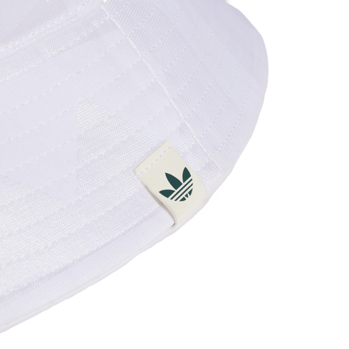 Close-up of the brim of a white bucket hat featuring a small off-white tag with a dark green Adidas Trefoil logo. Set against a solid black background.