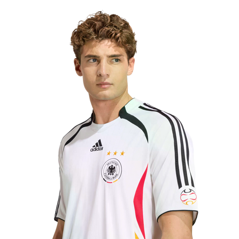 A man with curly hair wearing a white Adidas Germany national football team jersey, looking off to the side against a white background.