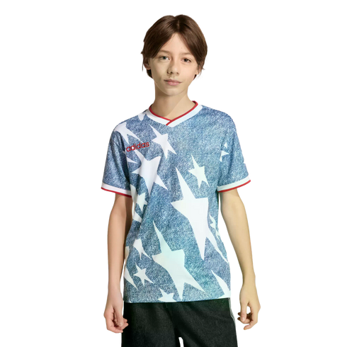 A young person wearing a blue and white star-patterned Adidas shirt with red trim and black shorts, standing against a black background.