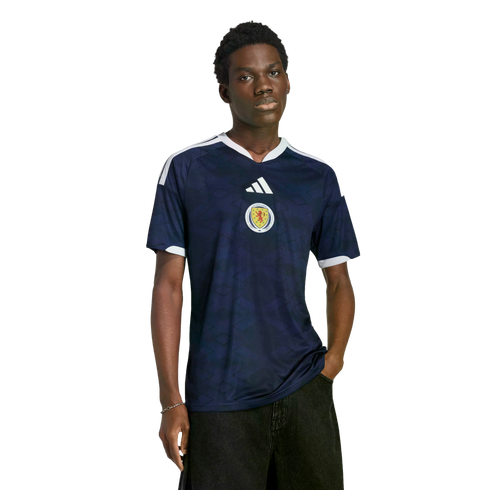 A young Black man models a navy blue Adidas Scotland soccer jersey against a solid black background.