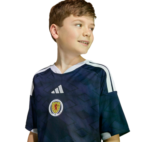 A smiling young boy wearing a navy blue Scotland soccer jersey with white stripes, looking to the side against a black background.
