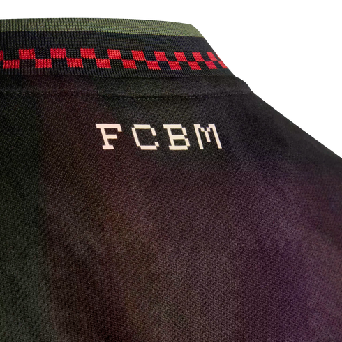 Close-up of a black jersey's back neck, showing white blocky "FCBM" text below a collar with green, navy, and red checkered patterns.