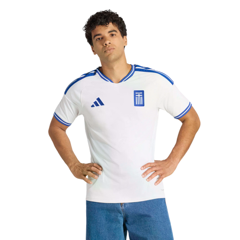 A man in a white Greece national team soccer jersey with blue trim stands with hands on his hips against a black background.