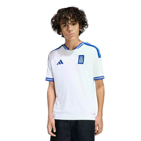A young person with curly dark hair wearing a white Greece national team soccer jersey with blue accents, standing against a solid black background.