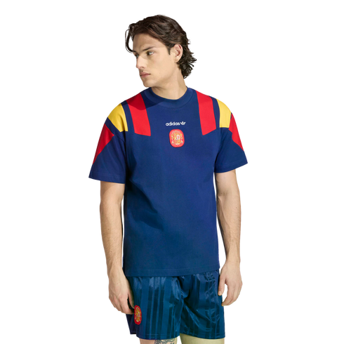 A man modeling a navy blue Adidas Spain football kit featuring a t-shirt with red and yellow shoulder panels and matching vertical-striped shorts.