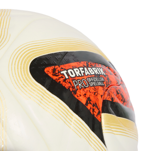 Close-up of a white soccer ball featuring gold curved lines and an orange and black graphic with the word "TORFABRIK."
