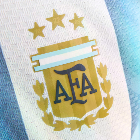 Close-up of the Argentina national football team crest on a blue and white striped jersey, featuring a gold emblem with "AFA" initials, laurel leaves, and three gold stars above.