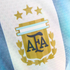 Close-up of the Argentina national football team crest on a blue and white striped jersey, featuring a gold emblem with "AFA" initials, laurel leaves, and three gold stars above.