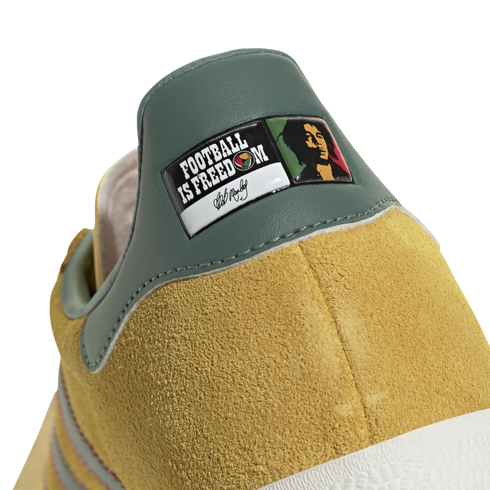 Close-up of a yellow suede shoe's green heel tab with a graphic patch reading "FOOTBALL IS FREEDOM," featuring Bob Marley’s portrait and signature.