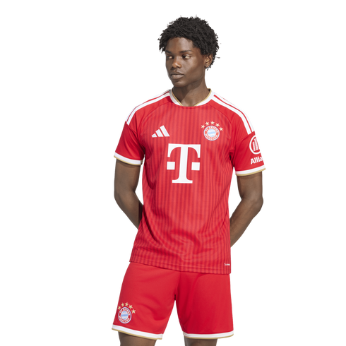 A man models a red and white FC Bayern Munich soccer kit. He stands against a white background, looking to his right with his hands behind his back.