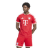 A man models a red and white FC Bayern Munich soccer kit. He stands against a white background, looking to his right with his hands behind his back.