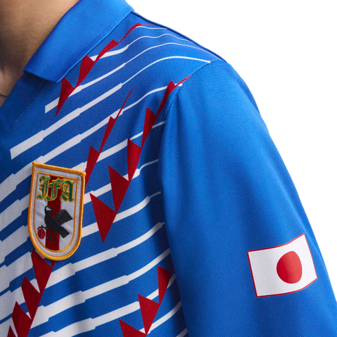 Close-up of a blue Japan national football jersey with white horizontal stripes, red jagged graphics, the JFA crest on the chest, and the Japanese flag on the sleeve.