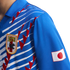 Close-up of a blue Japan national football jersey with white horizontal stripes, red jagged graphics, the JFA crest on the chest, and the Japanese flag on the sleeve.