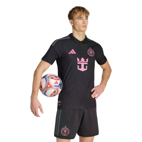 A male model wearing a black Inter Miami CF soccer kit with pink accents, holding a soccer ball under his arm against a plain black background.