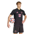A male model wearing a black Inter Miami CF soccer kit with pink accents, holding a soccer ball under his arm against a plain black background.