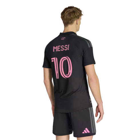 A person from behind wearing a black soccer jersey with the name "Messi" and the number "10" in pink on the back. He also wears matching black shorts with a pink Adidas logo on the leg, all set against a solid black background.