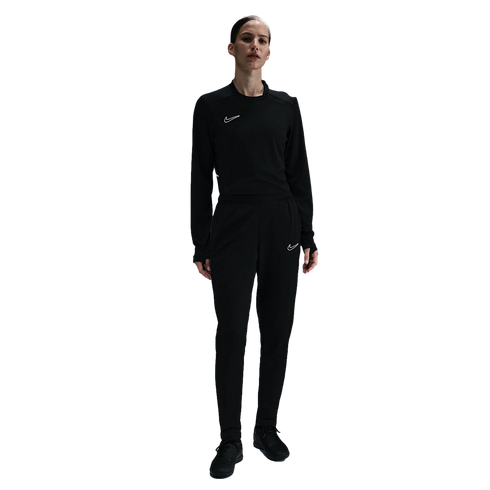 A woman standing in an all-black Nike tracksuit featuring a long-sleeve top and matching joggers, each with a small white swoosh logo, against a plain white background.