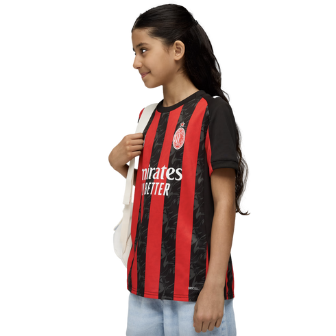 A young girl in a red and black AC Milan soccer jersey and light jeans smiles while carrying a white backpack against a white background.