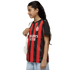 A young girl in a red and black AC Milan soccer jersey and light jeans smiles while carrying a white backpack against a white background.