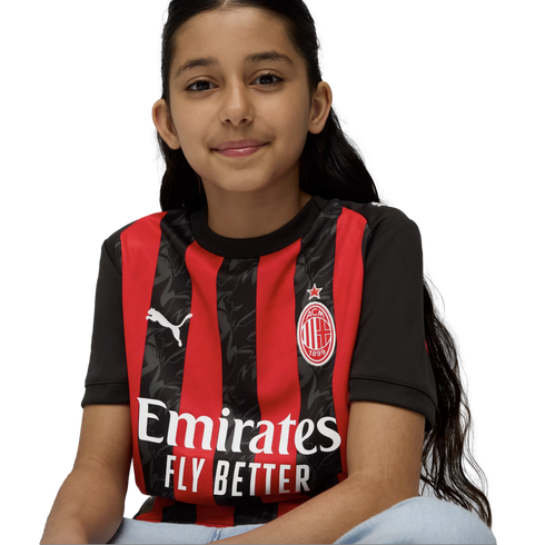 A smiling young girl with long dark hair sits against a white background wearing a red and black striped AC Milan soccer jersey.