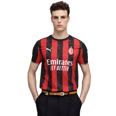 Young man wearing a red and black striped AC Milan soccer jersey, black trousers, and a tan belt, posing against a white background.