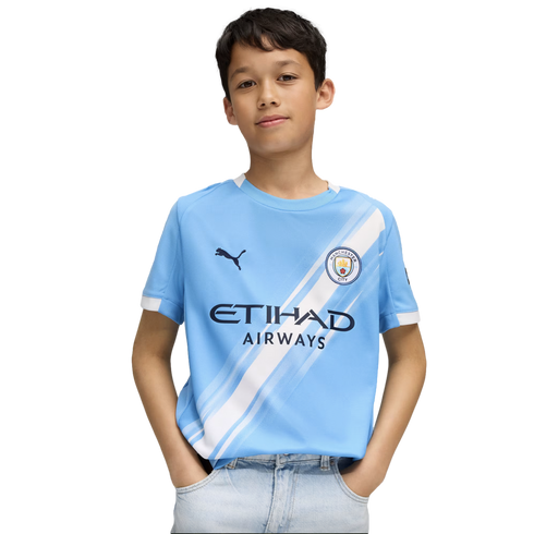 A young boy wearing a light blue Manchester City soccer jersey and light-wash jeans, standing with his hands in his pockets against a white background.