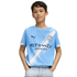 A young boy wearing a light blue Manchester City soccer jersey and light-wash jeans, standing with his hands in his pockets against a white background.