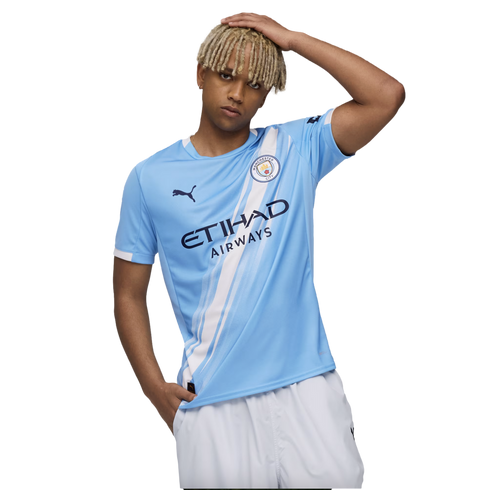 A young man with blonde dreadlocks wears a blue Manchester City soccer jersey and white shorts against a white background.