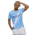 A young man with blonde dreadlocks wears a blue Manchester City soccer jersey and white shorts against a white background.