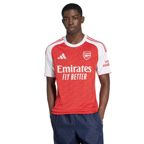 A man models the Arsenal home jersey, featuring a red body with white sleeves and sponsors. He is wearing dark blue shorts against a plain white background.