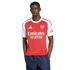 A man models the Arsenal home jersey, featuring a red body with white sleeves and sponsors. He is wearing dark blue shorts against a plain white background.
