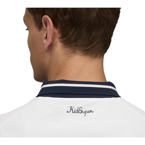 Close-up back view of a man's head and neck. He wears a white shirt with a navy striped collar and the "KidSuper" script logo printed on the upper back.