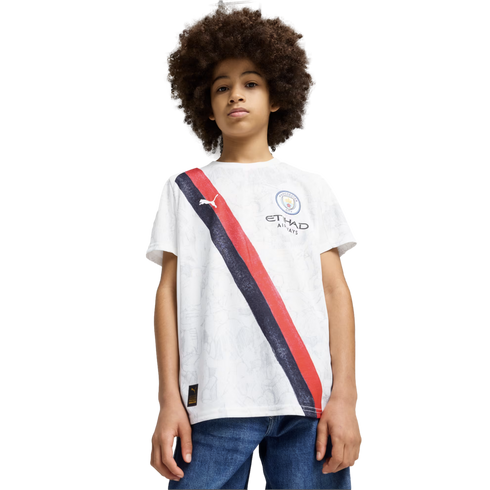 A boy with an afro wears a white Manchester City soccer jersey with a red and navy diagonal stripe, paired with blue jeans, against a plain white background.