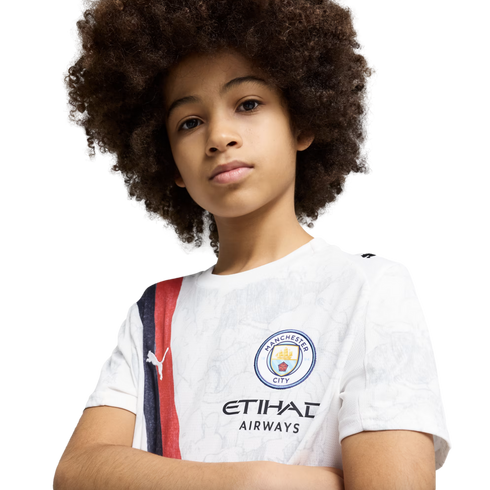 A young child with a large afro wears a white Manchester City soccer jersey, looking at the camera with arms crossed against a plain white background.