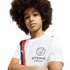 A young child with a large afro wears a white Manchester City soccer jersey, looking at the camera with arms crossed against a plain white background.