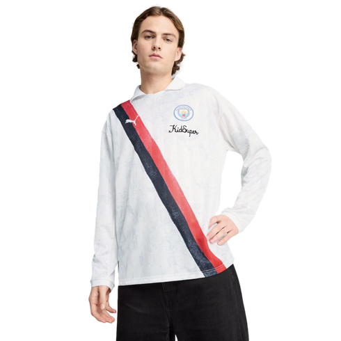 A man in a white Puma x KidSuper Manchester City long-sleeve polo featuring a red and navy sash, paired with black pants.