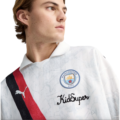 A young man wearing a white Manchester City x KidSuper Puma jersey featuring a red and black diagonal sash and the club crest.