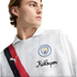 A young man wearing a white Manchester City x KidSuper Puma jersey featuring a red and black diagonal sash and the club crest.