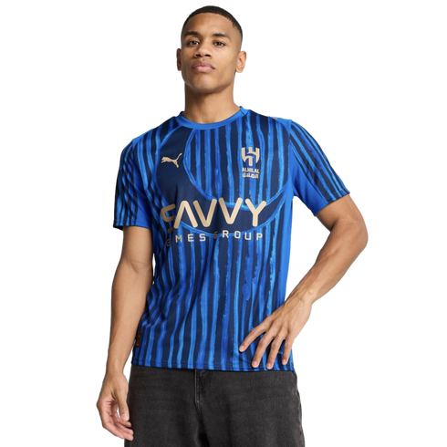 A man modeling an Al Hilal blue striped home soccer jersey with gold Puma and Savvy Games Group logos against a black background.