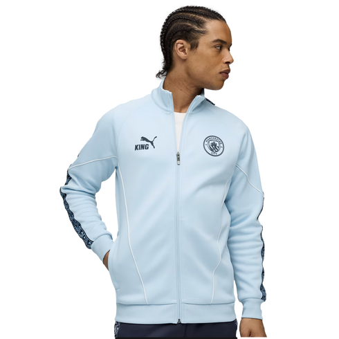 A man wearing a light blue Manchester City track jacket with the team crest, PUMA King logo, and patterned sleeve tape.